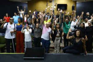 Grupo de pessoas diversas, incluindo pessoas com deficiência visual e mobilidade reduzida, reunidas em um auditório. Todas estão com as mãos levantadas, sorrindo e celebrando, demonstrando entusiasmo e união. Ao fundo, observa-se equipamentos de gravação e uma tela, indicando um evento formal. No primeiro plano, uma mulher sentada sorri, destacando-se pela expressão de alegria.